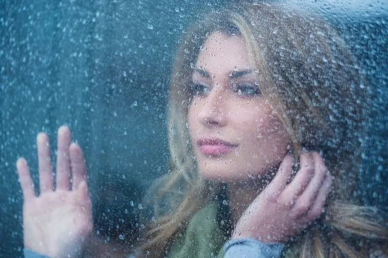 thoughtful young woman looking thru the glass window with raindrops
