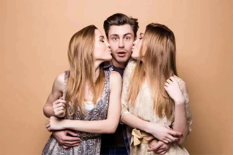 two women kissing one man hugging them both with a surprise expression on his face