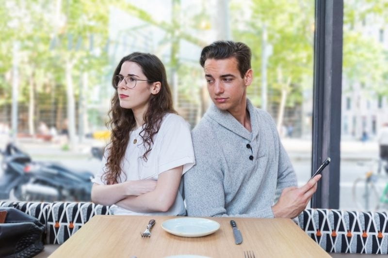 donna sconvolta in un ristorante, seduta accanto al suo fidanzato con in mano un cellulare
