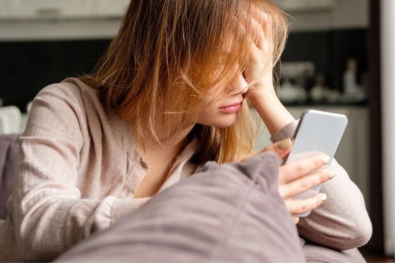 upset woman leaning on the couch looking at her smartphone inside living room