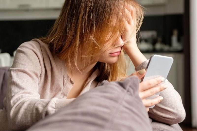 mulher perturbada, debruçada no sofá, a olhar para o seu smartphone na sala de estar