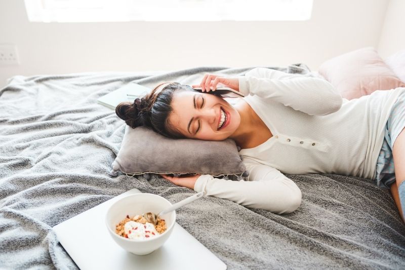 woman answering phone while lying in bed with food beside her