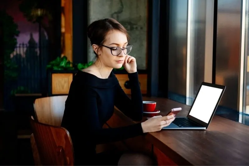 woman checking mobile phone while sitting at the table with laptop while working 