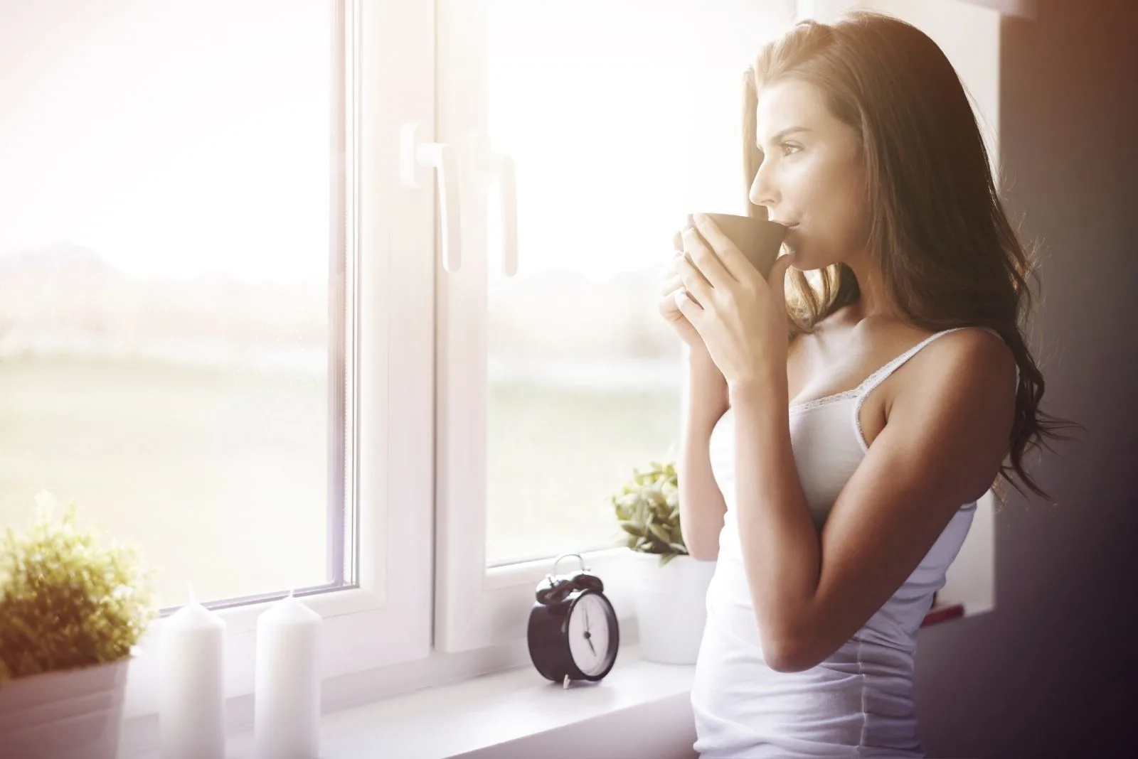 woman drinking coffee looking outside the window with an alarm clock nearby