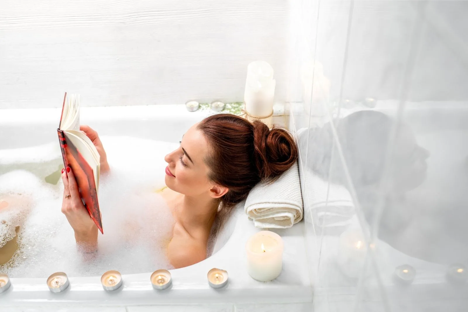 woman enjoyed bathing in the tub with lighted candles around while reading a book