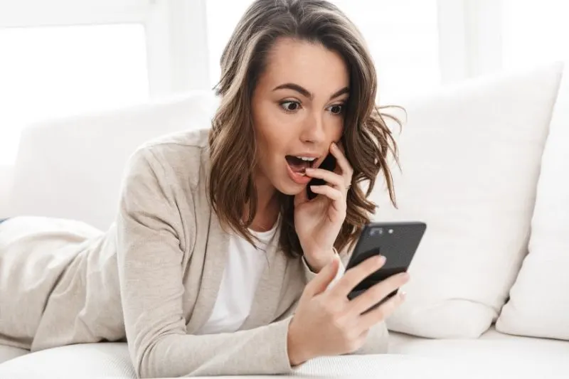 woman excited seeing the message on her smartphone while lying on the sofa inside living room