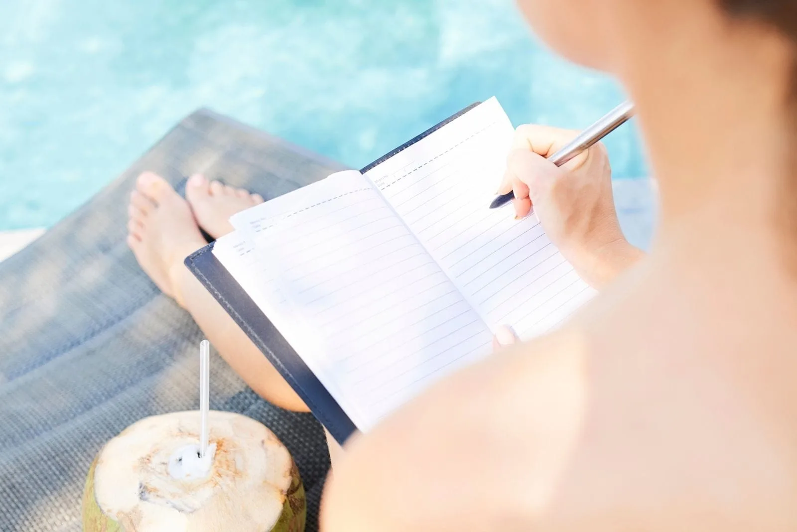 woman filling gratitude journal at the poolside with a coconut drink beside her