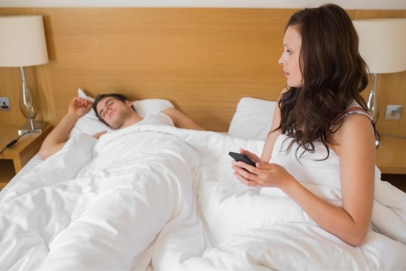 woman holding mobile phone while man sleeping on bed