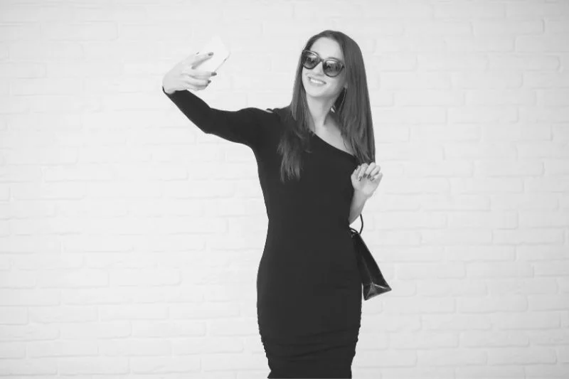 woman in black dress taking a selfie standing near a white brick wall