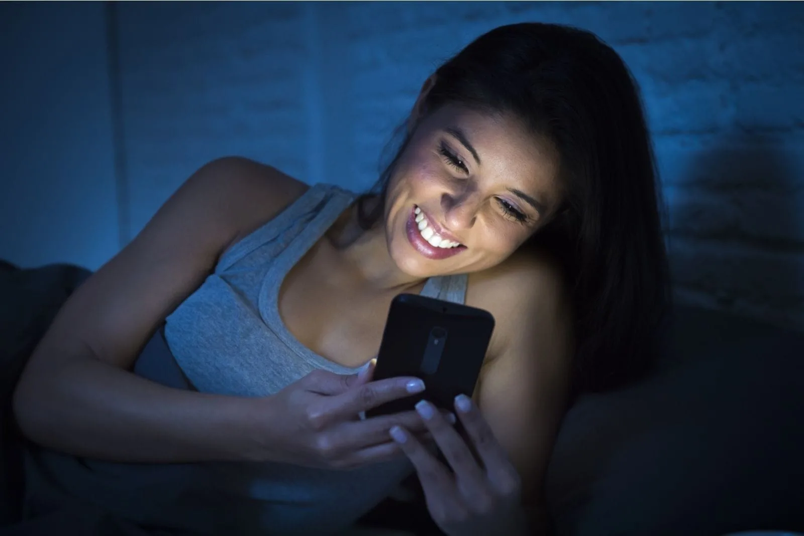 woman in tank top smiling while looking at her smartphone in bed with lights off