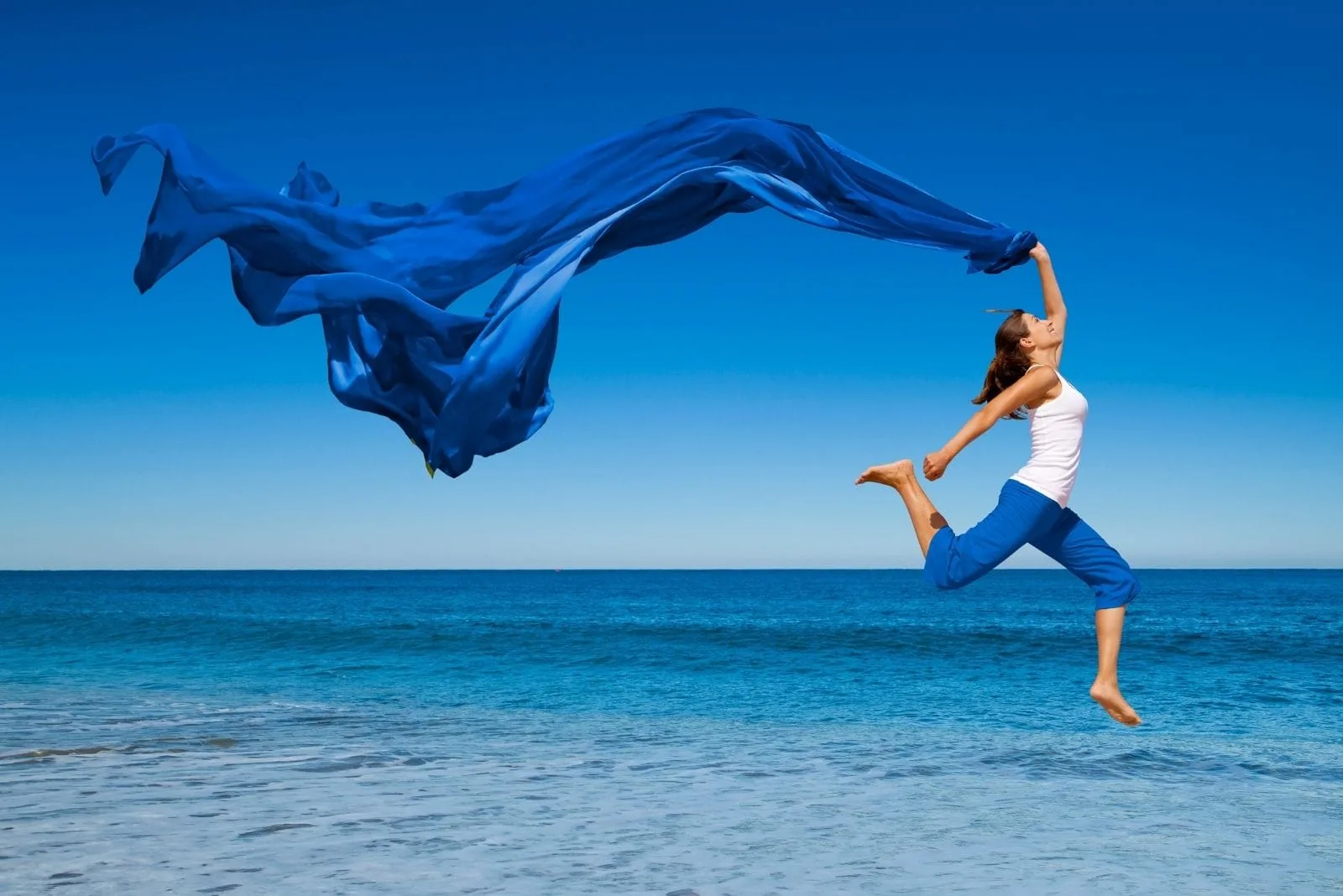 woman jump with joy near the blue sea carrying a blue cloth blown by the wind