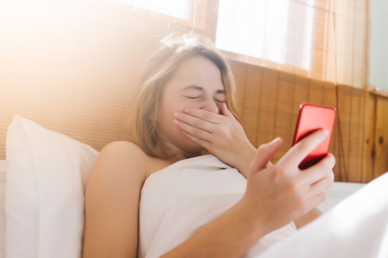 woman laying in bed sneezing while holding her smartphone