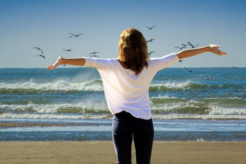 woman on beach spreading her arms facing the sea with a number of birds flying above