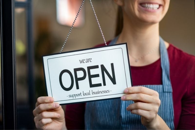 mujer abriendo un nuevo negocio sosteniendo un sinage "OPEN" y sonriendo en imagen recortada