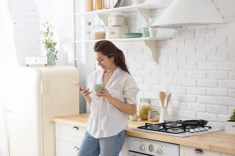 woman smiling and standing in the kitchen holding a mu and reading a text message from her cellphone