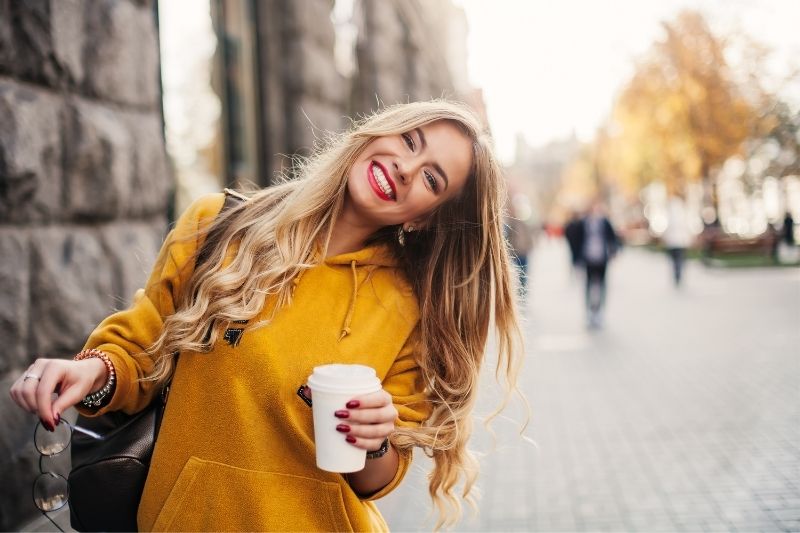 mulher a sorrir na rua, segurando uma chávena de café e inclinando a cabeça