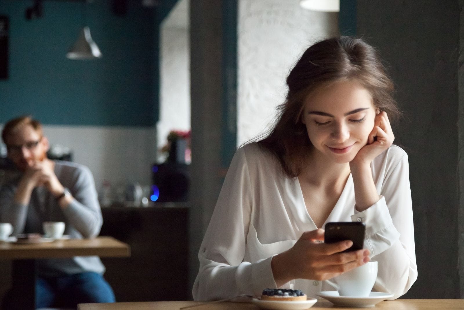 donna che fissa il suo smartphone sorridendo all'interno di un caffè con un uomo in fondo che la fissa