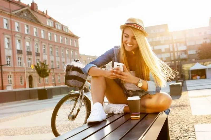 woman texting outdoors while sitting on a bench with coffee and a bicycle
