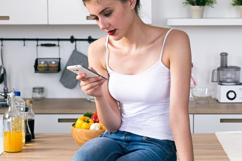 mujer usando el móvil sentada en la encimera de madera de la cocina