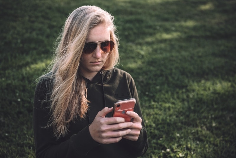 Mulher loira a utilizar o smartphone enquanto está sentada na relva