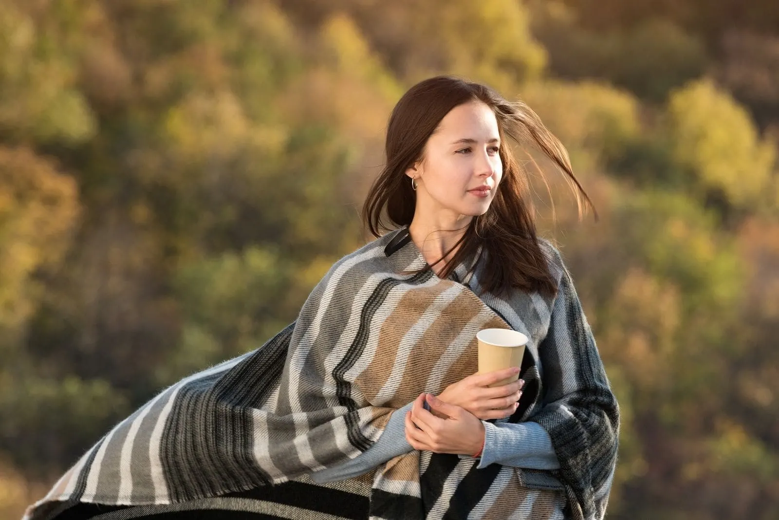 woman wrapped with blanket walking outdoors carrying coffee in paper cup