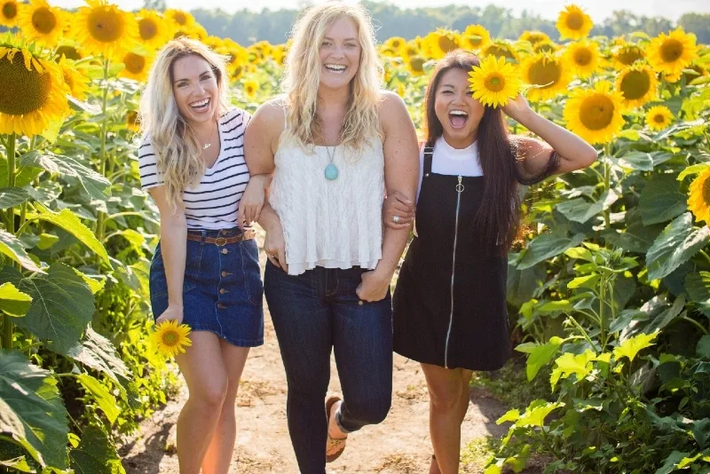 three women holding hands while standing between sunflowers