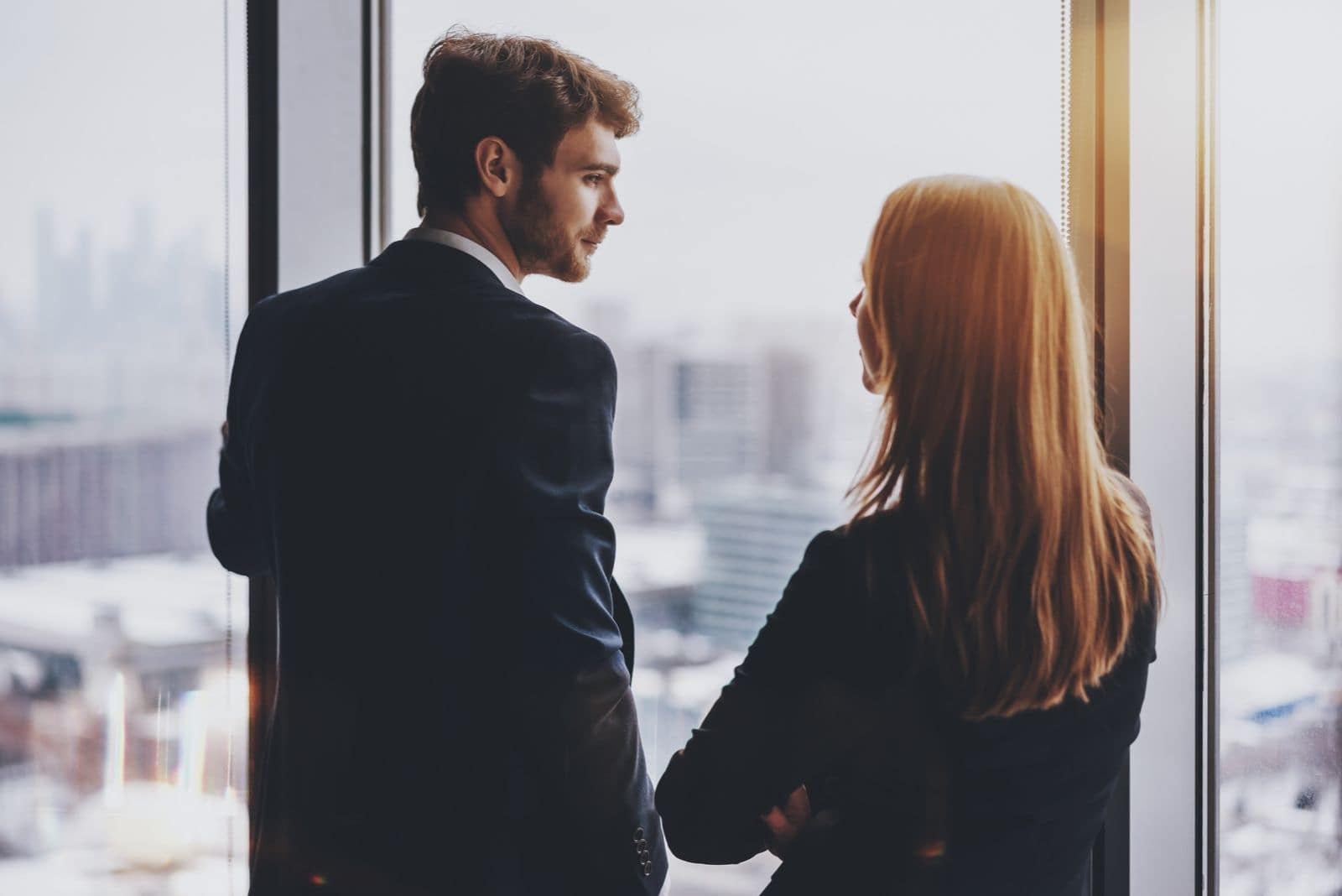young bearded business man talking to his female colleague seriously while standing near the glass windows of the office facing the city