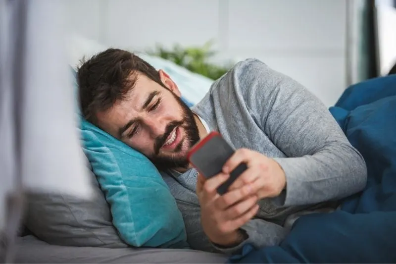young bearded man lying in bed and texting 
