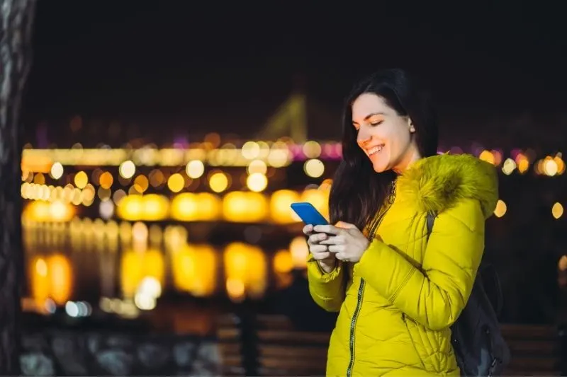 young caucasian woman texting outdoors in the night with city lights at the background