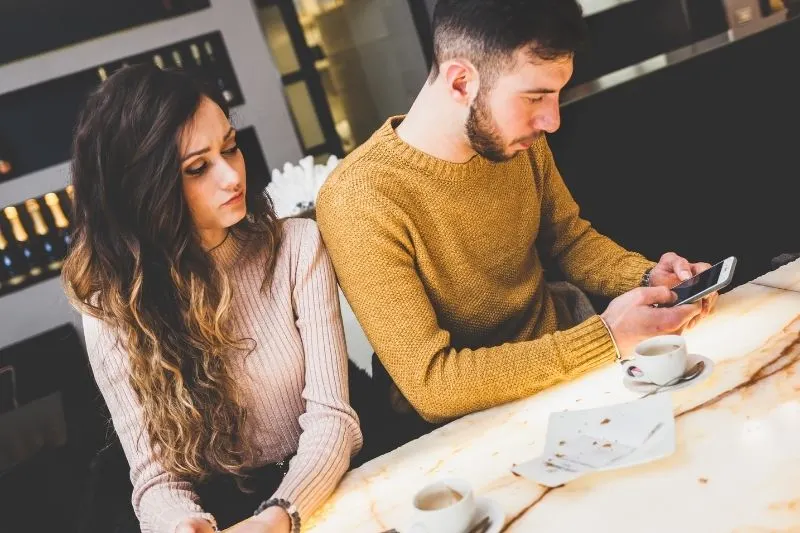young couple at a cafe not talking to each other with man at cellphone