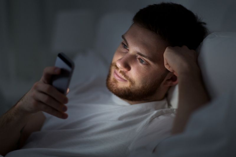giovane uomo affascinante che legge i messaggi sul suo cellulare sdraiato sul letto