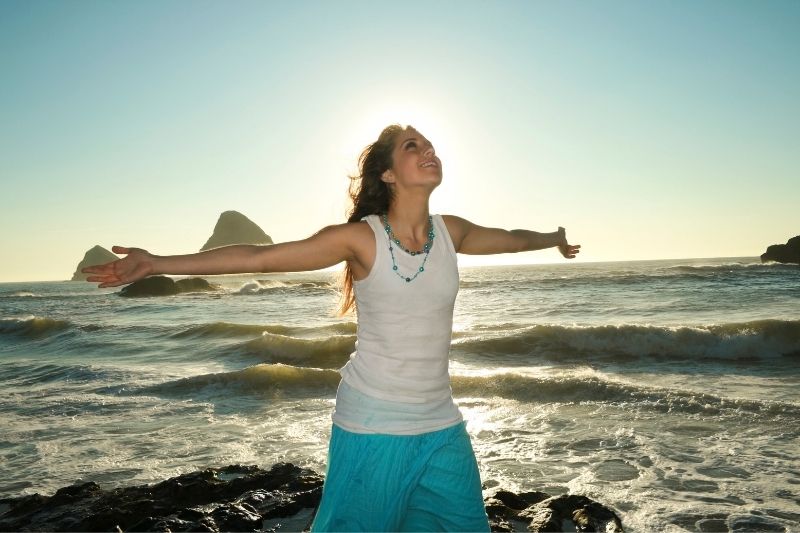 young inspired woman standing with arms raised near the sea 