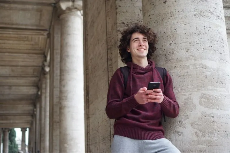 young man leaningon a circular column of a building texting and smiling while thinking