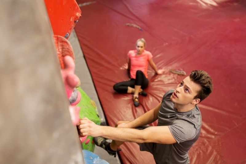 hombre joven escalada en roca dentro de gimnasio y una mujer en un lejano sentado en ángulo superior