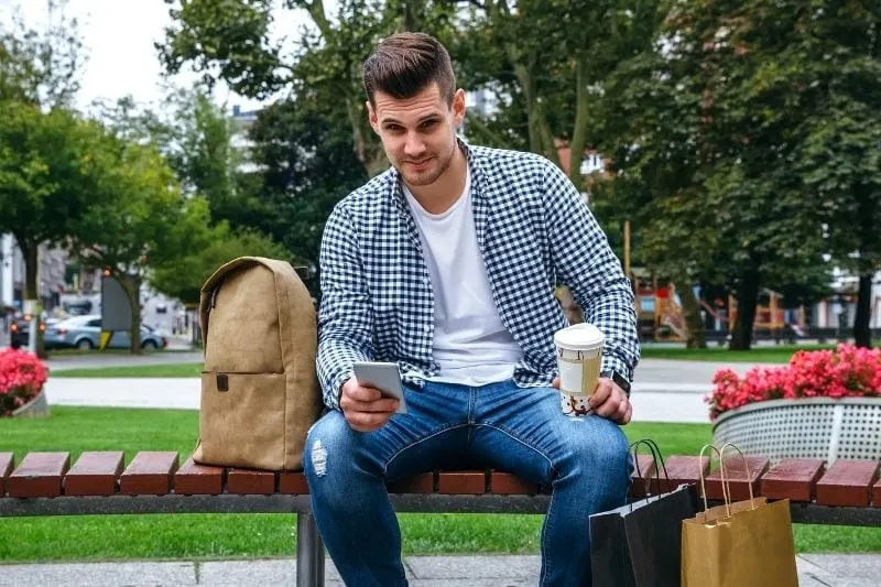 young man smiling at the camera holding coffee cup sitting on the park bench with shoppings bags on the ground