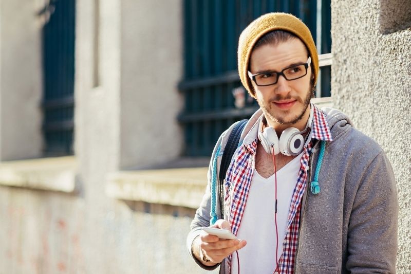 young man with bonnet texting while walking with headphone around his neck looking away