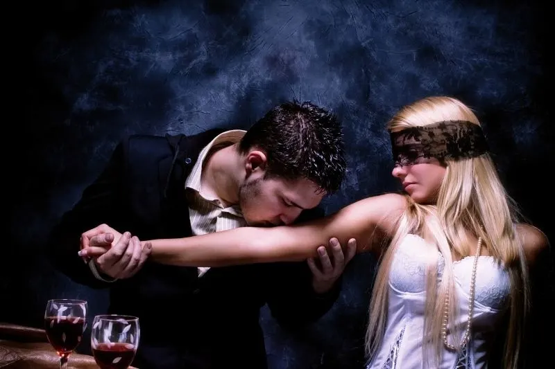 young woman covered her eyes with black lace with arms kissed by a man in suit 