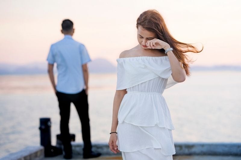 young woman crying wearing white dress walking away from a guy facing a body of water