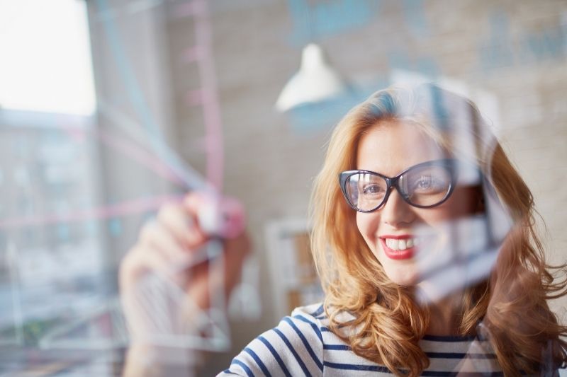 giovane donna che disegna sul vetro sorridendo con gli occhiali da vista