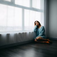 donna triste in maglione blu seduta sul pavimento vicino alla finestra