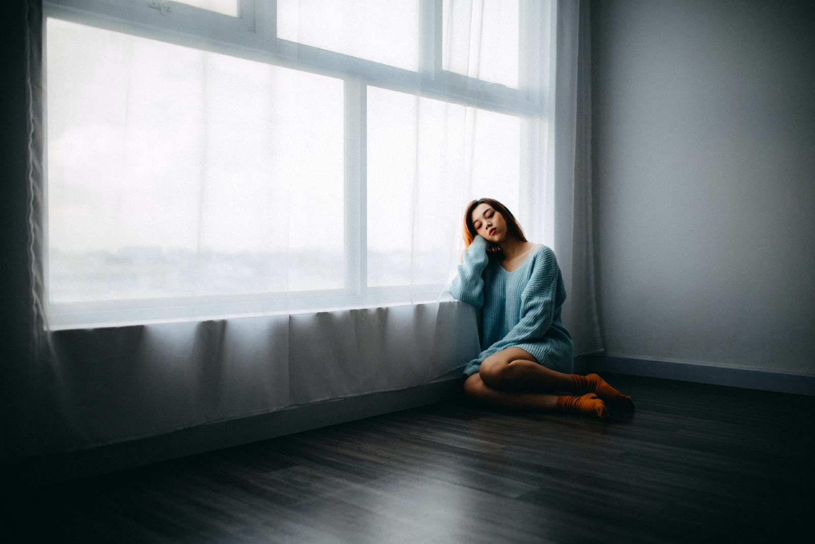 sad woman in blue sweater sitting on floor near window