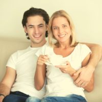 young man's arm around the older woman's shoulder while drinking coffee and sitting in the sofa inside living room