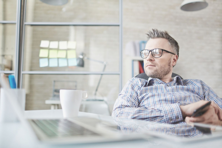 pensive young man at the office holding his phone sitting by his office table looking away