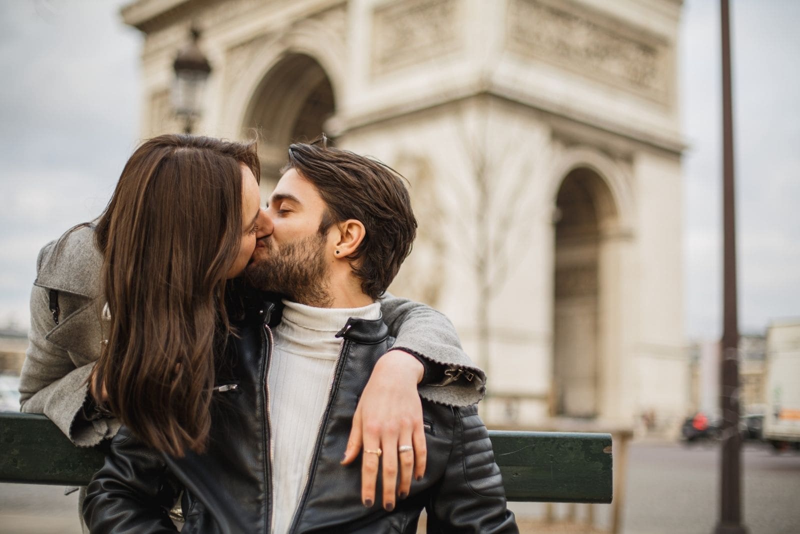 Parisian man and woman are french kissing near the triumphal arch with the man sitting on the bench