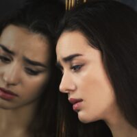a sad woman with long black hair leaning against the mirror