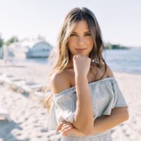 mulher com um vestido sem ombros, de pé na praia, a olhar para a câmara e a sorrir com os braços cruzados