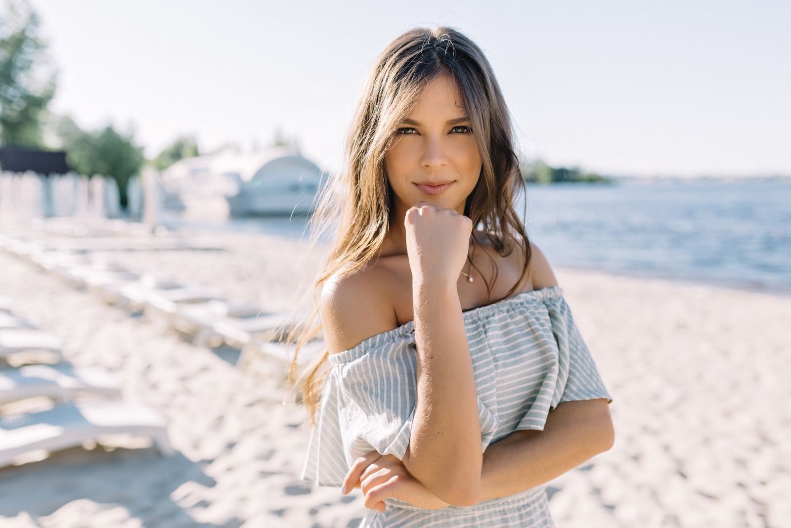 woman wearing an off shoulder dress standing in the beach looking at the camera smiling with crossed arms