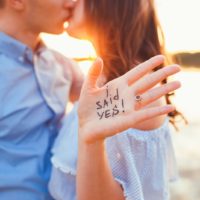 woman showing her hand with words i said yes kissing with a man in cropped image during sunset