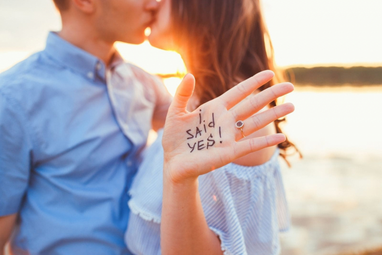 mulher mostrando a sua mão com as palavras "eu disse sim" beijando-se com um homem numa imagem recortada durante o pôr do sol