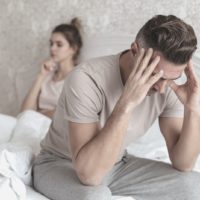 sad man sitting in the bed with his girlfriend sitting at the background inside the bedroom